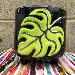 May include: Black ceramic planter with a green and silver monstera leaf design. The planter has three short legs.