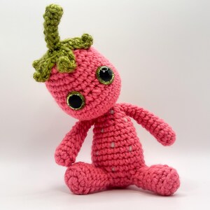May include: A pink crocheted strawberry plush toy with green leaves and black eyes. The toy is sitting on a white surface.