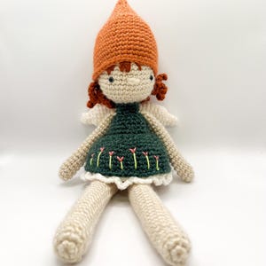 Op de afbeelding: Een gehaakte pop met een groene jurk, een oranje hoed en witte vleugels. De pop heeft een wit gezicht met geborduurde ogen en rood garen haar.