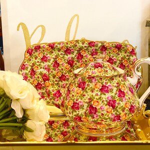 May include: A floral tea set with a teapot and serving tray. The set features a pattern of red, pink, and orange roses on a cream background. A bouquet of white roses sits in front of the tea set. The word "Rejoice" is written in gold.