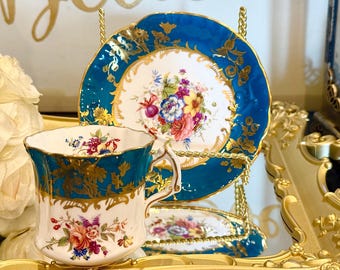 May include: A vintage teacup and saucer set with a floral design. The cup and saucer have a white base with colorful flowers and gold accents. The rims are a deep teal blue. The set is displayed on a gold-colored stand.