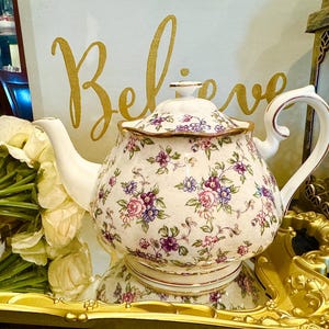 Könnte beinhalten: Eine weiße Porzellan-Teekanne mit einem Blumenmuster in Rosa, Lila und Blau. Die Teekanne hat einen gebogenen Griff und Ausguss sowie einen passenden Deckel. Das Wort "Believe" ist in goldener Schrift im Hintergrund geschrieben. Die Teekanne steht auf einem goldenen Tablett.