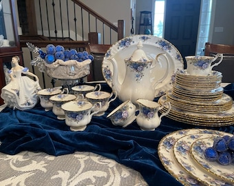 May include: A white tea set with a blue and gold floral pattern. The set includes a teapot, creamer, sugar bowl, cups, saucers, and a serving platter. The set is displayed on a blue velvet tablecloth with blue and white decorative balls.