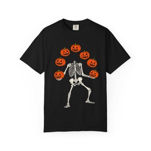 Op de afbeelding: Een zwart t-shirt met korte mouwen en een grafische print van een wit skelet dat jongleert met zes oranje pompoenen. Het skelet staat in een dynamische houding met de armen omhoog en de pompoenen zijn in een boog boven het skelet geplaatst.