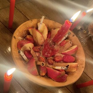 May include: A wooden bowl filled with sliced red apples, cinnamon sticks, and several lit cigarettes. A single pink candle is burning in the center of the bowl, surrounded by scattered brown powder. Additional red candles are placed outside the bowl.