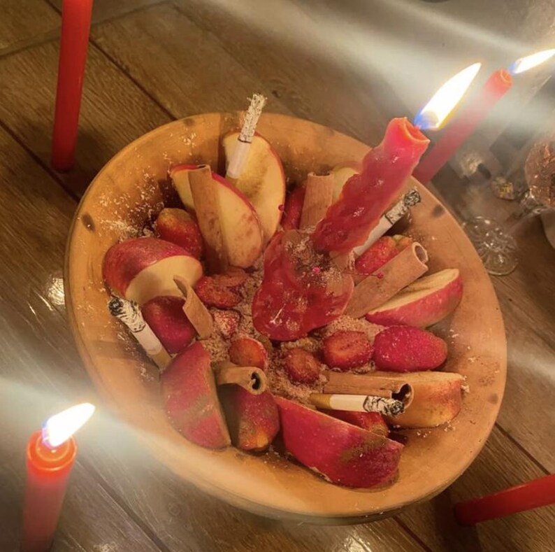 May include: A wooden bowl filled with sliced red apples, cinnamon sticks, and several lit cigarettes. A single pink candle is burning in the center of the bowl, surrounded by scattered brown powder. Additional red candles are placed outside the bowl.