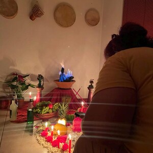 May include: A ritual altar setup featuring a central terracotta bowl with blue flames, surrounded by red candles, fresh fruit including grapes and a pineapple, green plants, and small figurines. The display is arranged on a red cloth.