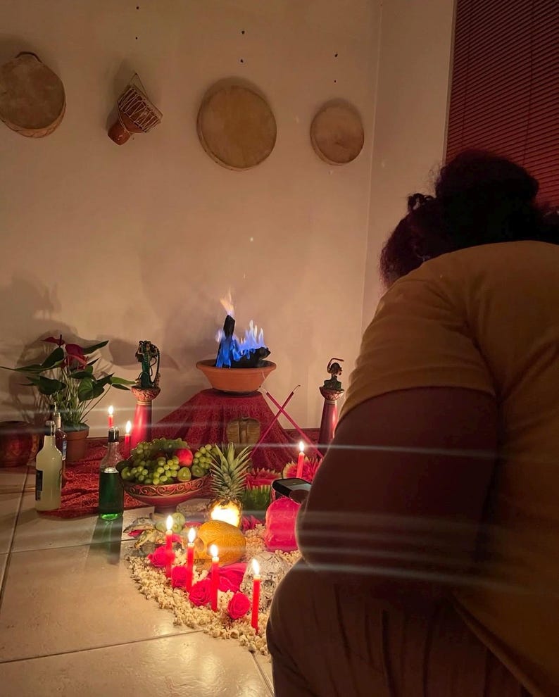 May include: A ritual altar setup featuring a central terracotta bowl with blue flames, surrounded by red candles, fresh fruit including grapes and a pineapple, green plants, and small figurines. The display is arranged on a red cloth.