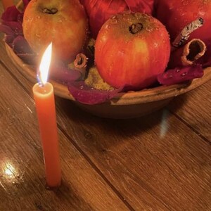 May include: A centerpiece featuring several red apples with water droplets, a red satin ribbon, and cinnamon sticks arranged in a bowl. A single orange taper candle with a lit flame stands upright in the foreground on a wooden surface.