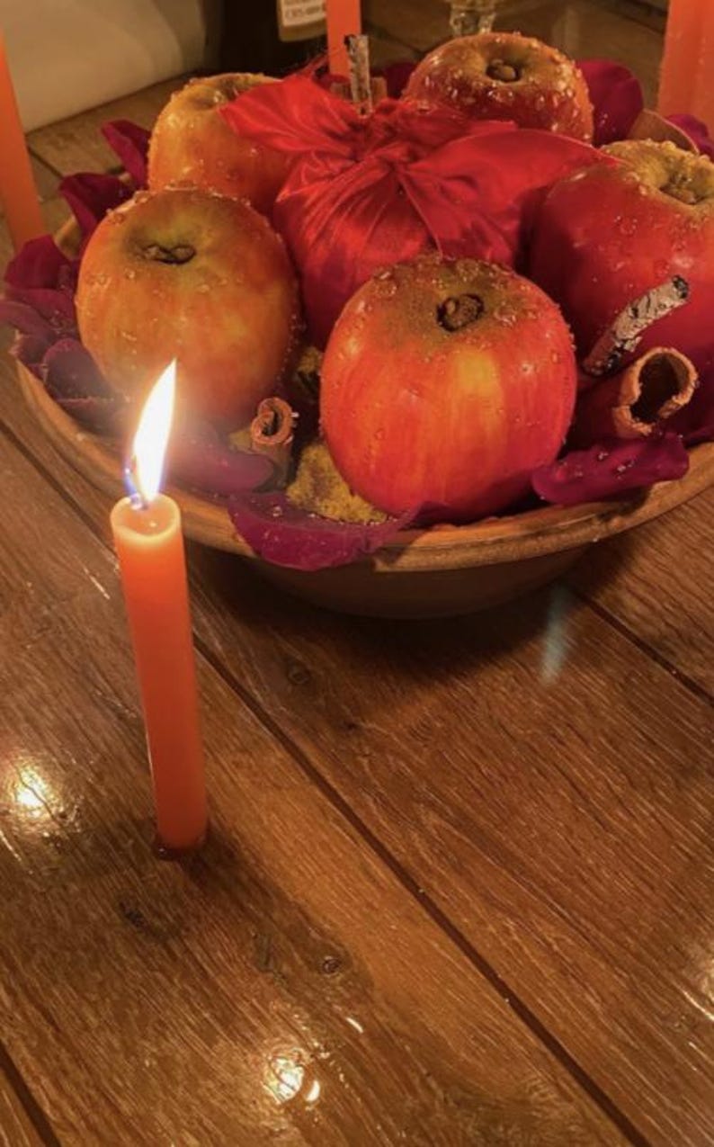 May include: A centerpiece featuring several red apples with water droplets, a red satin ribbon, and cinnamon sticks arranged in a bowl. A single orange taper candle with a lit flame stands upright in the foreground on a wooden surface.