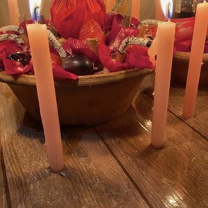 May include: A round brown bowl filled with red rose petals, dark grapes, strawberries, and cinnamon sticks, surrounded by several tall, thin, light pink taper candles with lit flames, all arranged on a dark wooden surface.