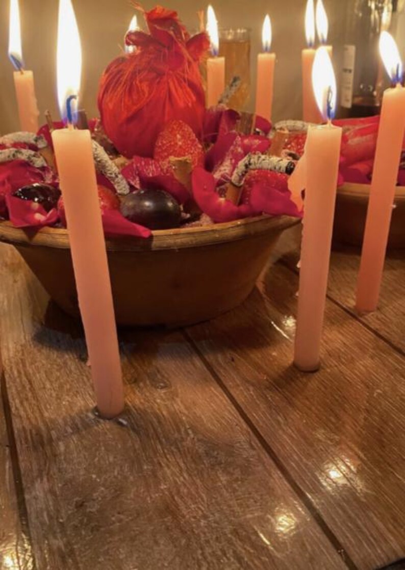 May include: A round brown bowl filled with red rose petals, dark grapes, strawberries, and cinnamon sticks, surrounded by several tall, thin, light pink taper candles with lit flames, all arranged on a dark wooden surface.