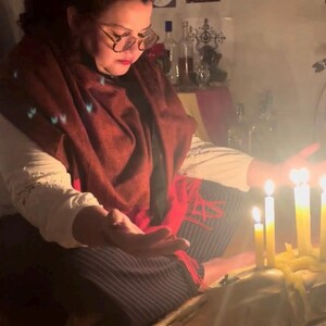 May include: A person wearing a white shirt, dark pinstriped pants, and a deep red shawl, sitting with hands outstretched toward several lit yellow candles placed on a large animal skull. The scene is dimly lit, highlighting the warm glow of the flames.