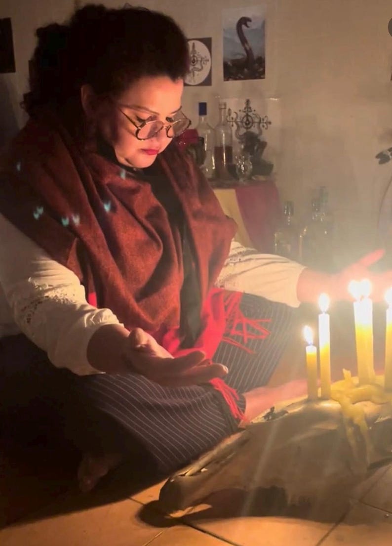 May include: A person wearing a white shirt, dark pinstriped pants, and a deep red shawl, sitting with hands outstretched toward several lit yellow candles placed on a large animal skull. The scene is dimly lit, highlighting the warm glow of the flames.