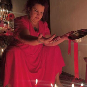 May include: A person wearing a vibrant red dress with short sleeves and a V-neckline. The person is holding a silver metal bowl and pouring a red liquid over their hand. Several red candles are lit in the foreground and background.