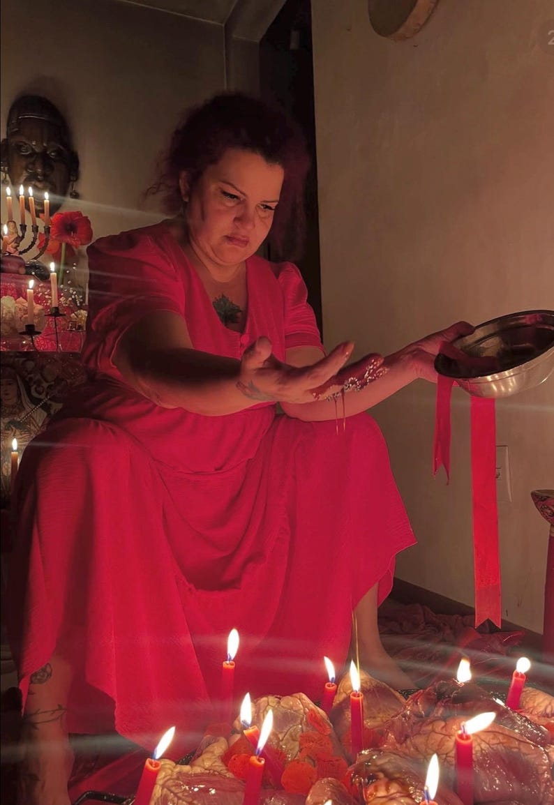 May include: A person wearing a vibrant red dress with short sleeves and a V-neckline. The person is holding a silver metal bowl and pouring a red liquid over their hand. Several red candles are lit in the foreground and background.