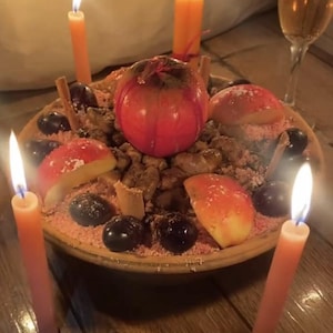 May include: A round ceramic bowl filled with a mixture of dark grapes, sliced red apples, and cinnamon sticks, topped with a whole red apple. Four lit orange taper candles surround the bowl, and a glass of amber liquid stands nearby.