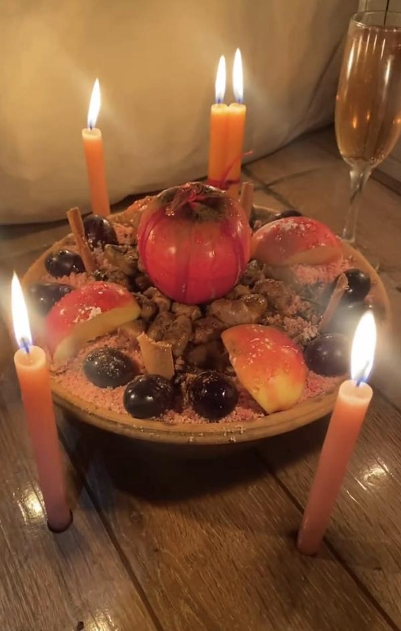 May include: A round ceramic bowl filled with a mixture of dark grapes, sliced red apples, and cinnamon sticks, topped with a whole red apple. Four lit orange taper candles surround the bowl, and a glass of amber liquid stands nearby.