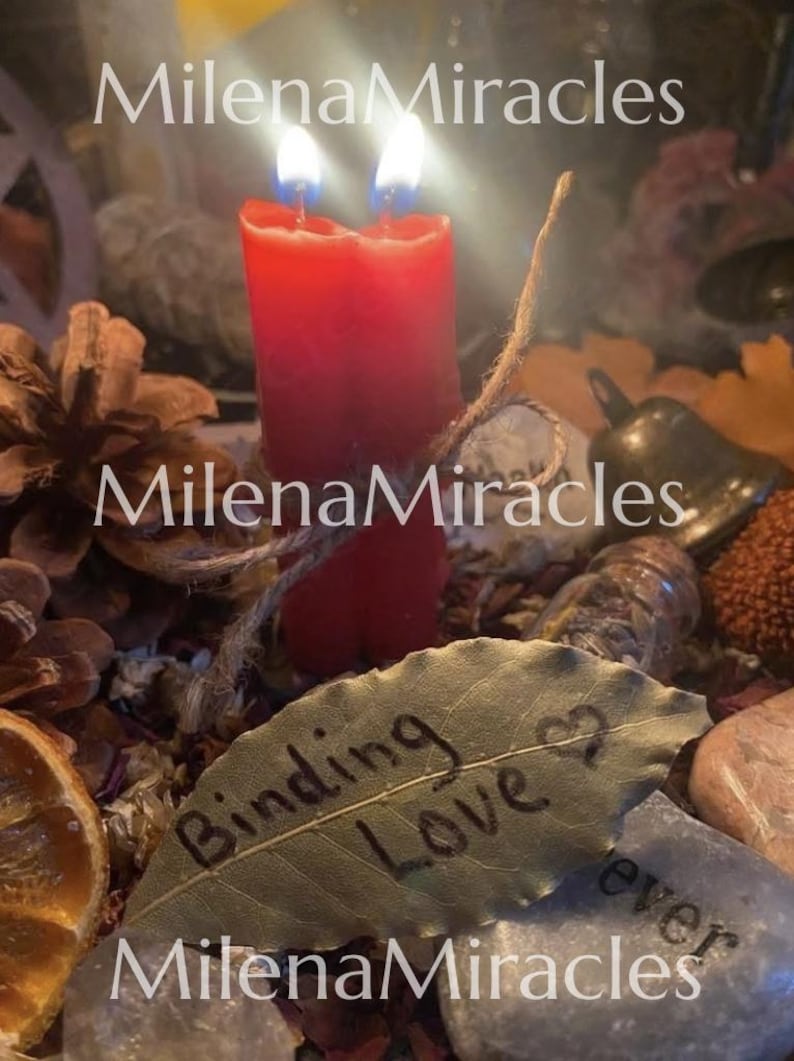 May include: Two red candles joined together with a twine string, positioned behind a dried leaf with the text Binding Love and a heart symbol written on it, surrounded by pinecones, a small metal bell, and various stones.