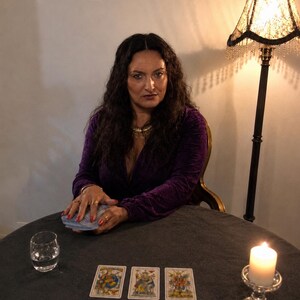 May include: A person with long dark hair wearing a deep purple velvet long-sleeve top, sitting at a round table with a deck of cards, a glass of water, and a lit white candle. Three tarot cards are laid out in a row on the dark gray tablecloth.