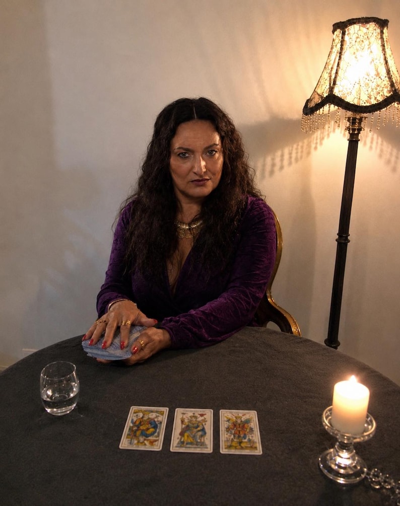 May include: A person with long dark hair wearing a deep purple velvet long-sleeve top, sitting at a round table with a deck of cards, a glass of water, and a lit white candle. Three tarot cards are laid out in a row on the dark gray tablecloth.
