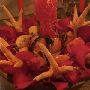 May include: A centerpiece featuring a central red candle, surrounded by red rose petals, sliced apples, and several light-colored chicken feet. Two additional red candles stand in the background, with a glass of liquid visible behind them.