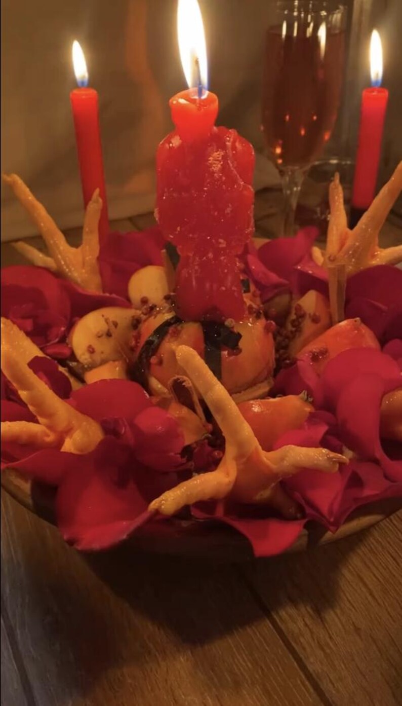 May include: A centerpiece featuring a central red candle, surrounded by red rose petals, sliced apples, and several light-colored chicken feet. Two additional red candles stand in the background, with a glass of liquid visible behind them.