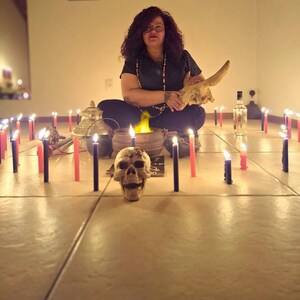 May include: A human skull sits in the foreground surrounded by numerous lit red and black candles arranged on a tiled floor. Behind the skull, a small cauldron with a yellow flame, a metallic lidded container, and a clear glass bottle are visible.