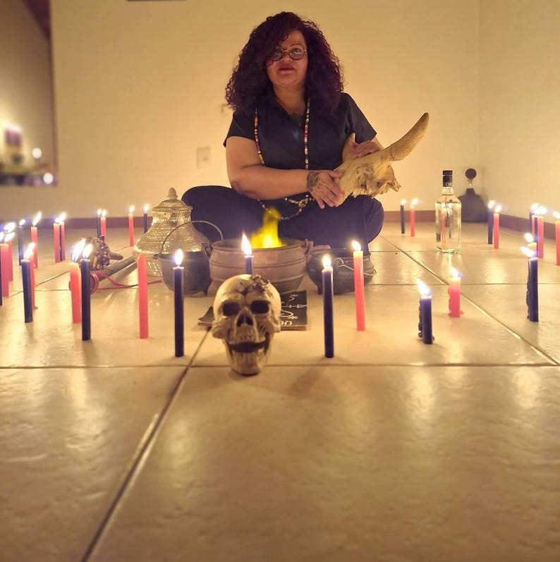 May include: A human skull sits in the foreground surrounded by numerous lit red and black candles arranged on a tiled floor. Behind the skull, a small cauldron with a yellow flame, a metallic lidded container, and a clear glass bottle are visible.