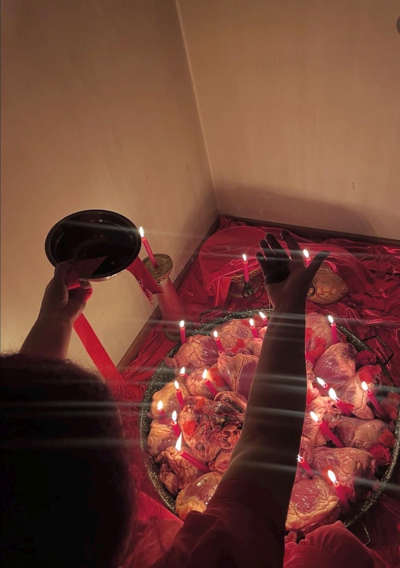 May include: A large metal pan filled with multiple raw animal hearts arranged in a circle, illuminated by numerous thin red candles. The scene is set against a red fabric background, with a small metal bowl held in one hand.