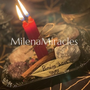 May include: Two red candles with lit flames are tied together with twine, resting on a black tray alongside raw crystals, a cinnamon stick, and a bay leaf inscribed with the text Binding Love and three small heart symbols.