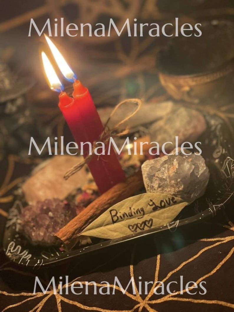 May include: Two red candles with lit flames are tied together with twine, resting on a black tray alongside raw crystals, a cinnamon stick, and a bay leaf inscribed with the text Binding Love and three small heart symbols.