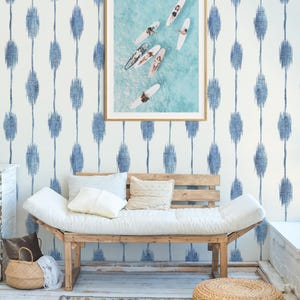 Pu&ograve; includere: Una fotografia incorniciata di surfisti su tavole da surf nell'oceano &egrave; appesa sopra una panca di legno con cuscini e cuscini bianchi. La parete &egrave; decorata con un motivo ikat blu e bianco. Un cesto intrecciato e un tappeto sono sul pavimento.