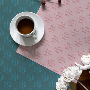 May include: A pink wrapping paper with the text "Your Artwork Here" repeated in a pattern. A cup of coffee and a bouquet of white flowers are on top of the paper.