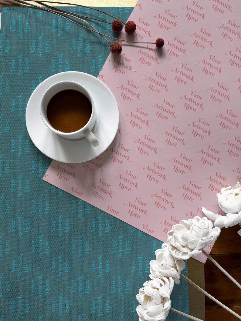 May include: A pink wrapping paper with the text "Your Artwork Here" repeated in a pattern. A cup of coffee and a bouquet of white flowers are on top of the paper.