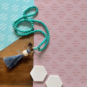 May include: A turquoise beaded necklace with a tassel charm and two white hexagonal tiles on a wooden surface. The tiles are on top of pink and blue patterned paper.