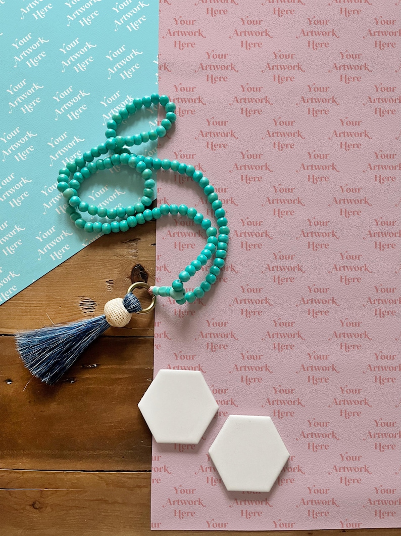 May include: A turquoise beaded necklace with a tassel charm and two white hexagonal tiles on a wooden surface. The tiles are on top of pink and blue patterned paper.