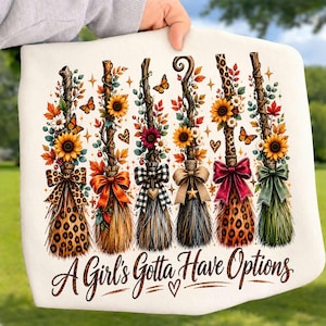 May include: A white sweatshirt featuring a graphic of six decorative brooms adorned with sunflowers, autumn leaves, butterflies, and various bows. The text "A Girl's Gotta Have Options" is printed in a dark brown script font at the bottom.
