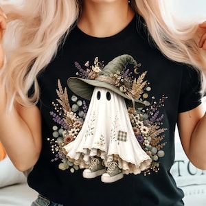 Peut inclure: Un t-shirt noir avec un motif représentant un fantôme blanc portant un chapeau vert olive à larges bords. Le fantôme est entouré de fleurs séchées et de blé, et porte des baskets vert olive. Il présente un petit empiècement à carreaux.