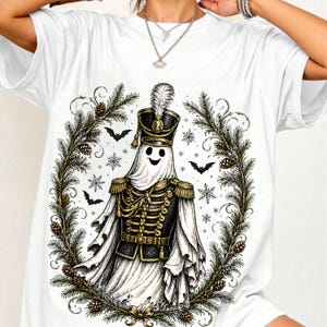 May include: A white short-sleeve t-shirt featuring a graphic print of a ghost dressed in a black and gold military-style marching band uniform with a feathered hat, surrounded by a wreath of pine branches, pinecones, snowflakes, and bats.