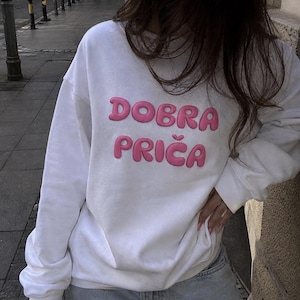 May include: A white long-sleeved sweatshirt featuring the text DOBRA PRIČA in raised, bubble-style pink lettering across the chest. The garment has a crew neckline, long sleeves with ribbed cuffs, and a relaxed fit, providing a clean look.