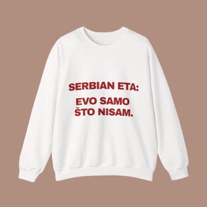 Pode incluir: Uma camisola branca de manga comprida com gola redonda e o texto vermelho escuro SERBIAN ETA: EVO SAMO ŠTO NISAM. impresso na frente. A peça tem gola, punhos e bainha canelados, com um corte padrão e uma cor branca sólida em toda a peça.