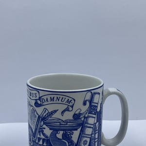 Op de afbeelding: Een witte keramische mok met een blauw en wit ontwerp met een griffin, boeken en de tekst "DAMNUM".