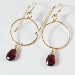 Puede incluir: Aretes de aro de oro con una piedra preciosa roja colgante. Los aros son texturizados y las piedras preciosas son facetadas.