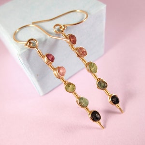 May include: A pair of gold earrings with a dangling design featuring five gemstones in shades of pink, green, and black.