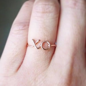 May include: A rose gold ring with the letters "XO" intertwined. The ring is worn on a finger.