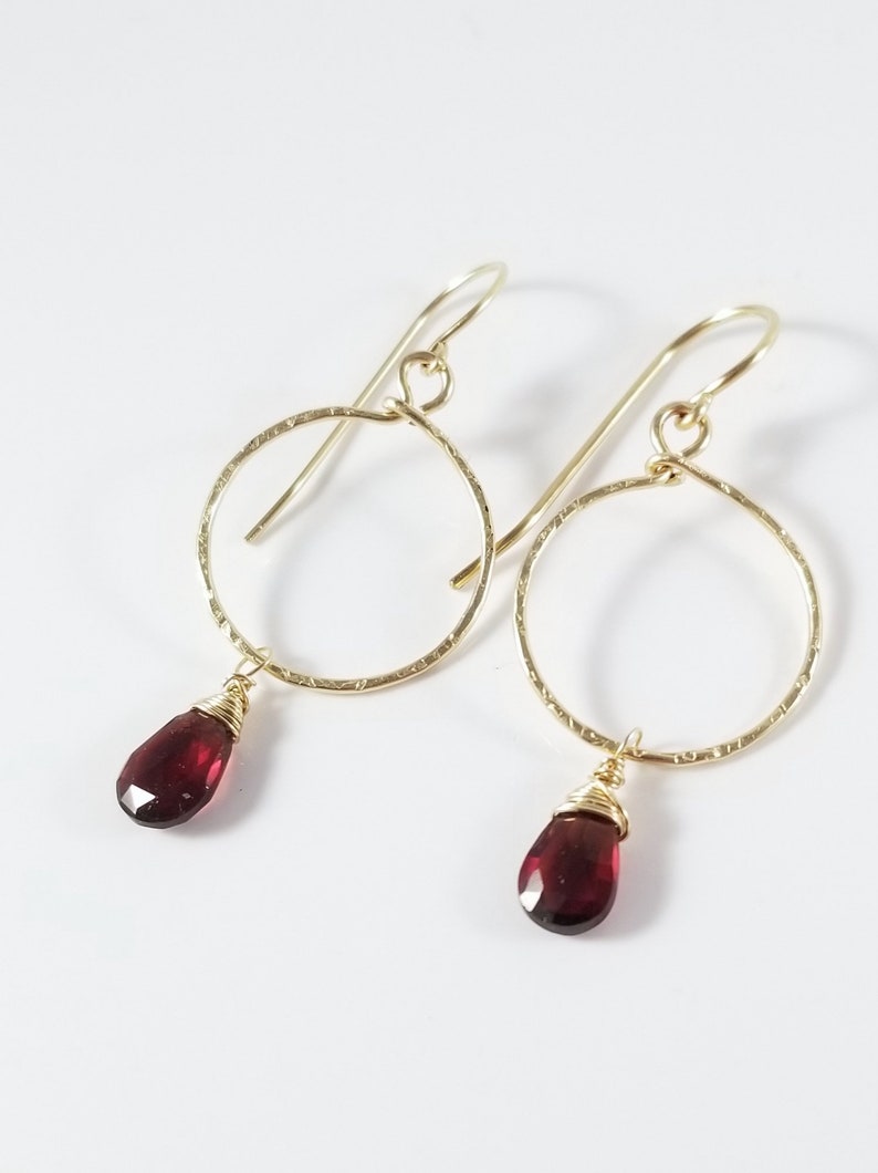 Puede incluir: Aretes de aro de oro con una piedra preciosa roja colgante. Los aros son texturados y las piedras preciosas son facetadas.