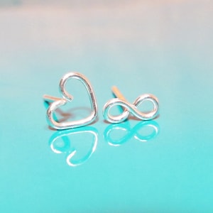 May include: A pair of silver earrings, one shaped like a heart and the other shaped like an infinity symbol. The earrings are on a turquoise background.
