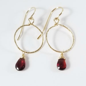 Puede incluir: Aretes de aro de oro con una piedra preciosa roja colgante. Los aros son texturizados y las piedras preciosas son facetadas.
