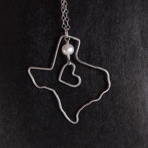 May include: A silver necklace with a Texas state outline pendant and a small pearl charm. The pendant also features a heart shape inside the state outline.